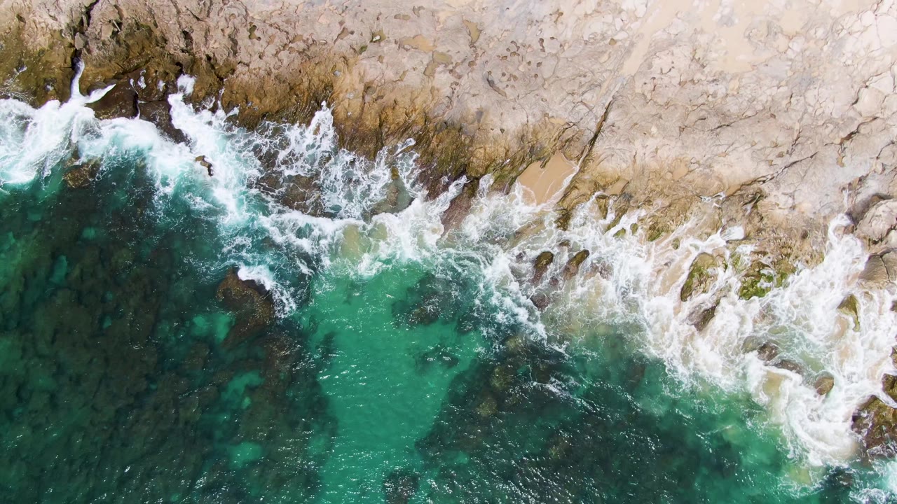 At Punta Gorda Beach in Baja California Sur, white foam spreads broadly across the upper rock edge while deeper teal water darkens the lower left of the overhead frame, with no visible people.