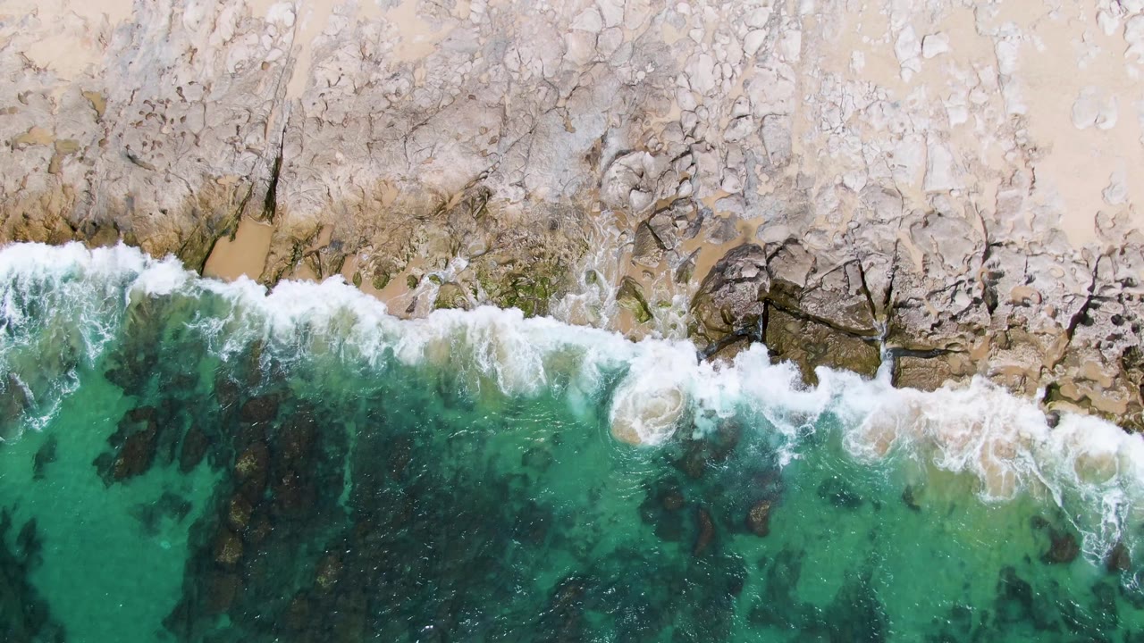 Overhead view at Punta Gorda Beach, Baja California Sur, where turquoise water meets a pale rocky shoreline with a thin band of white foam and no visible people.