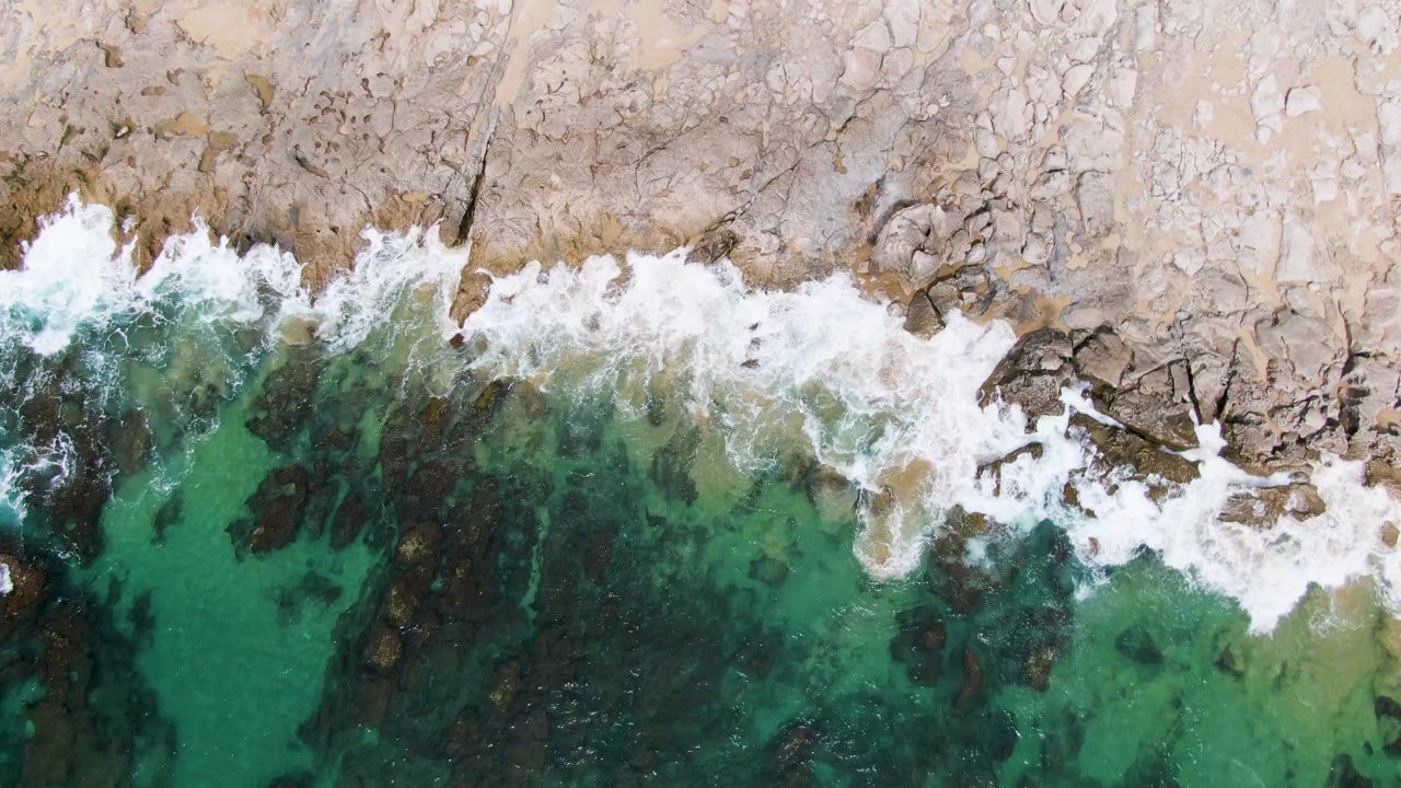 Overhead frame from Punta Gorda Beach, Baja California Sur, with a larger burst of white foam pushing onto jagged pale rocks above clear turquoise water and no visible people.