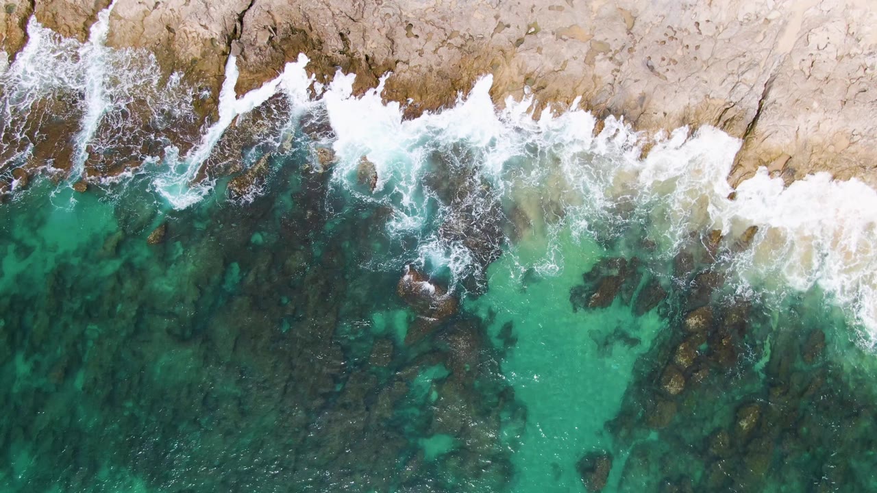 Aerial frame from Punta Gorda Beach showing a wider band of white wave foam over uneven tan rock, with clear green-blue water and dark underwater stones below, and no visible people.