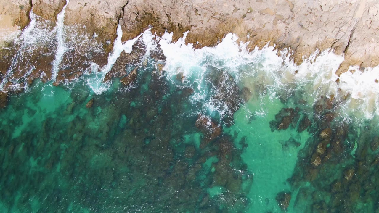 Top-down Punta Gorda Beach view with broken white surf along the upper rock edge and translucent turquoise water revealing dark submerged rock shapes below, with no visible people.