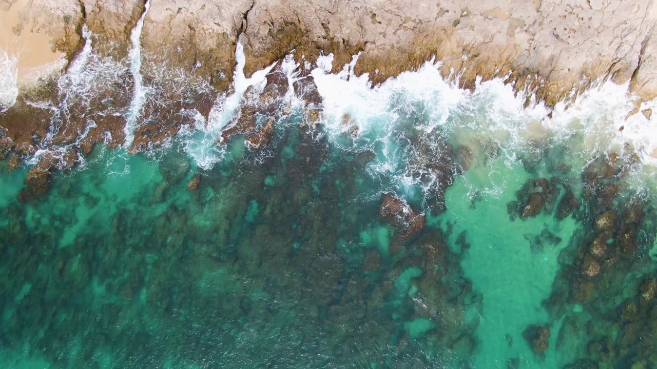 Overhead frame at Punta Gorda Beach where white foam clings to the tan rock boundary while darker green water and scattered underwater rocks fill the lower area, with no visible people.