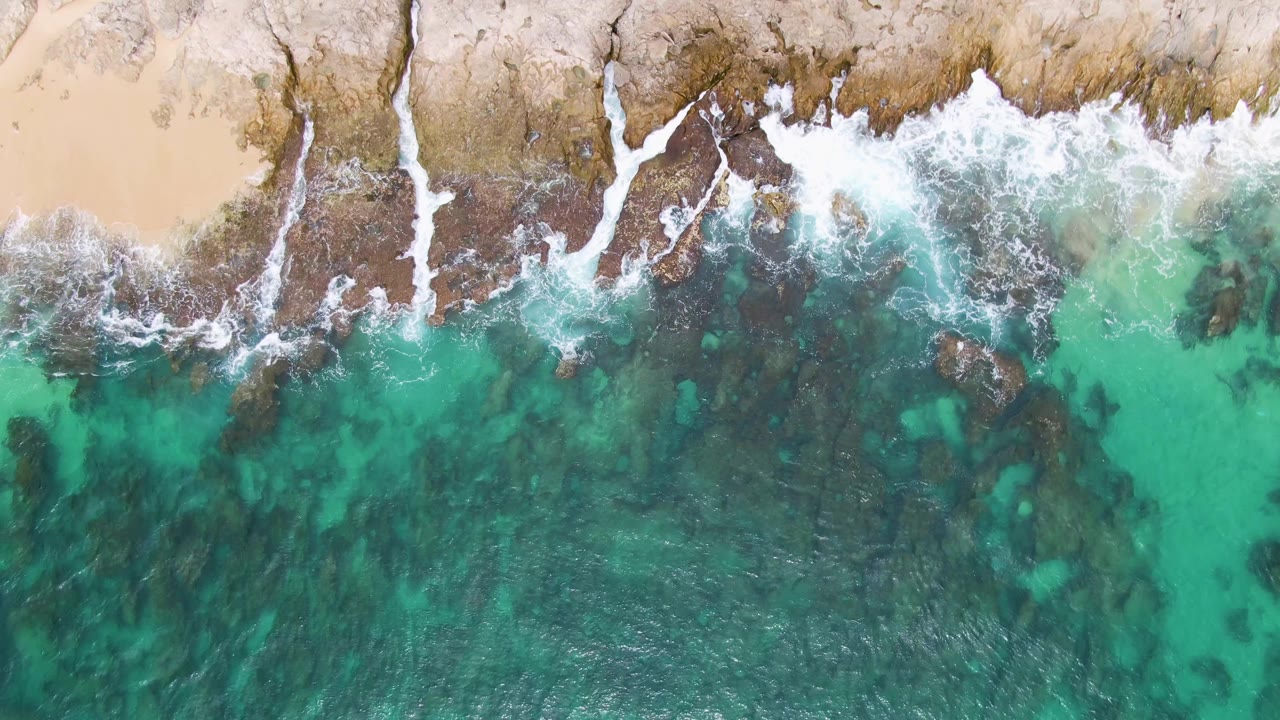 Overhead view at Punta Gorda Beach, Baja California Sur, with turquoise water meeting dark rocks and pale sand along the upper edge, no visible people.