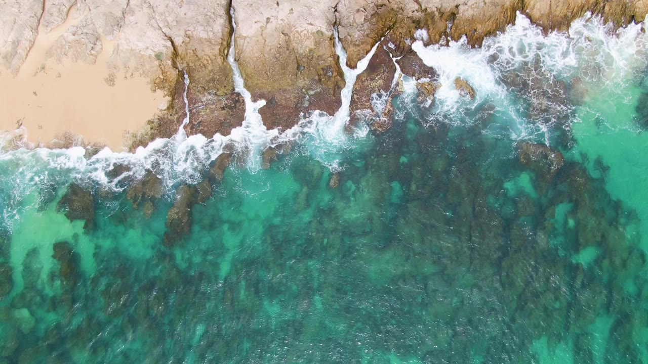 Over Punta Gorda Beach, the foamy wave line runs across the rocky shoreline with pale sand above and mottled turquoise water below, no visible people.