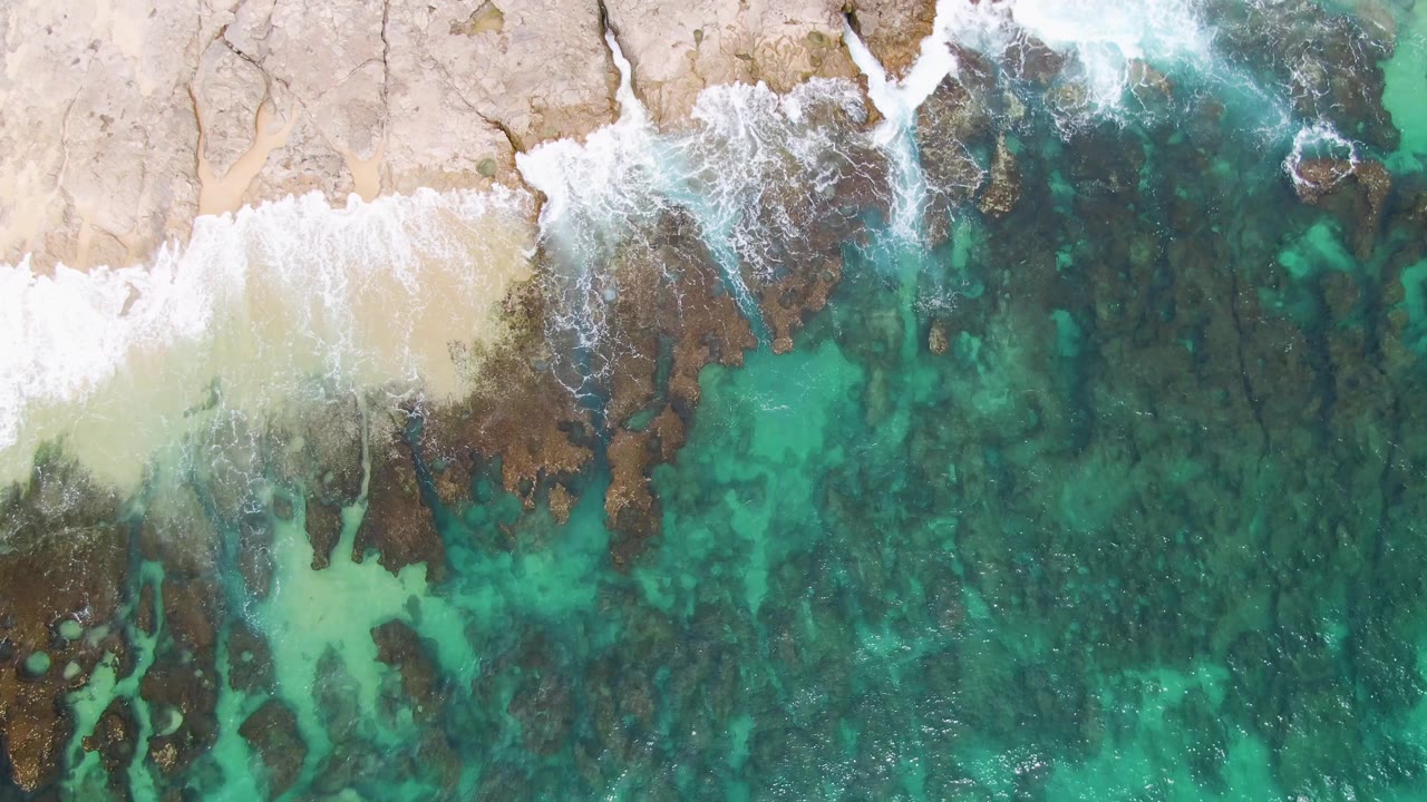 Overhead view of Punta Gorda Beach with pale sand, dark shoreline rocks, turquoise shallow water, white wave foam, and no visible people at the opening frame.