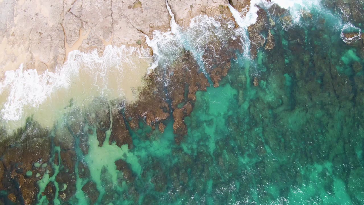 Aerial frame of Punta Gorda Beach where white foam spreads farther along the rocks, with sand above and clear green-blue water below, and no visible people.