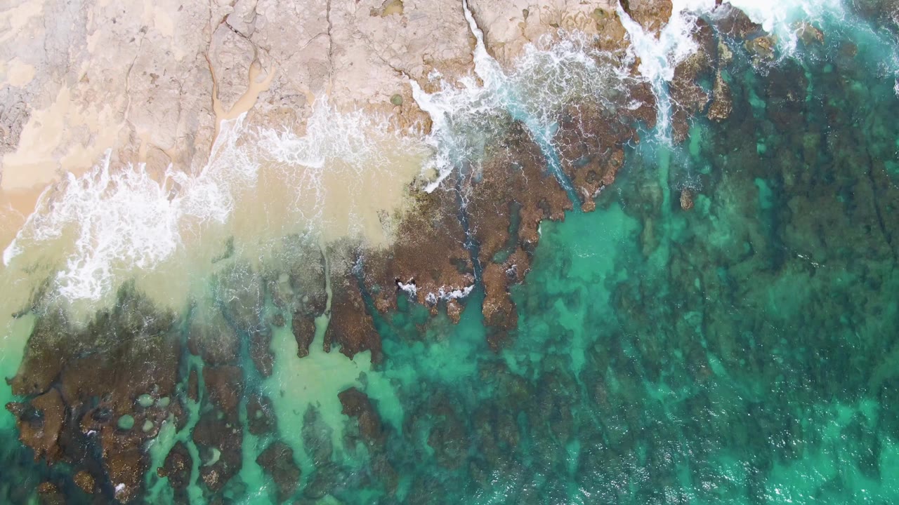 Overhead Punta Gorda Beach view with a broader band of sand, broken white surf on rock shelves, clear turquoise shallows, and no visible people.