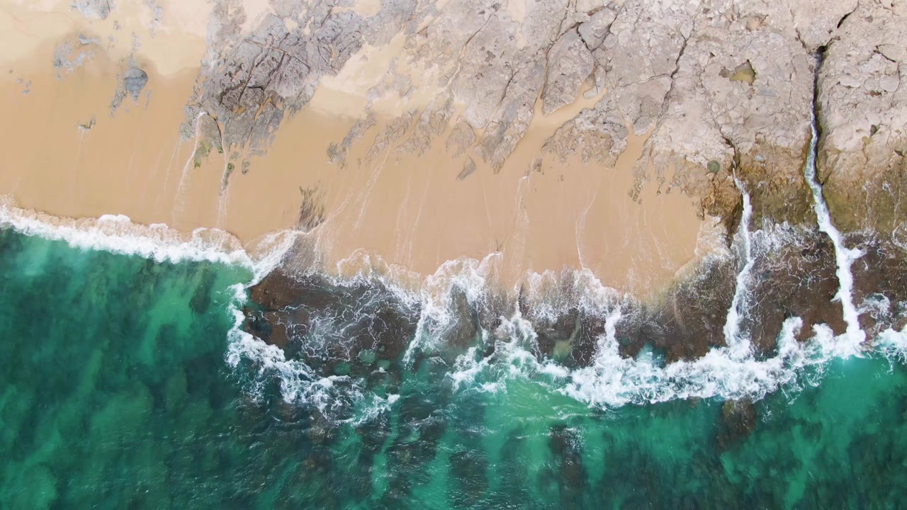 Aerial view of Punta Gorda Beach with a curved band of white surf between pale sand, gray rock, and deep turquoise water, with no visible people.
