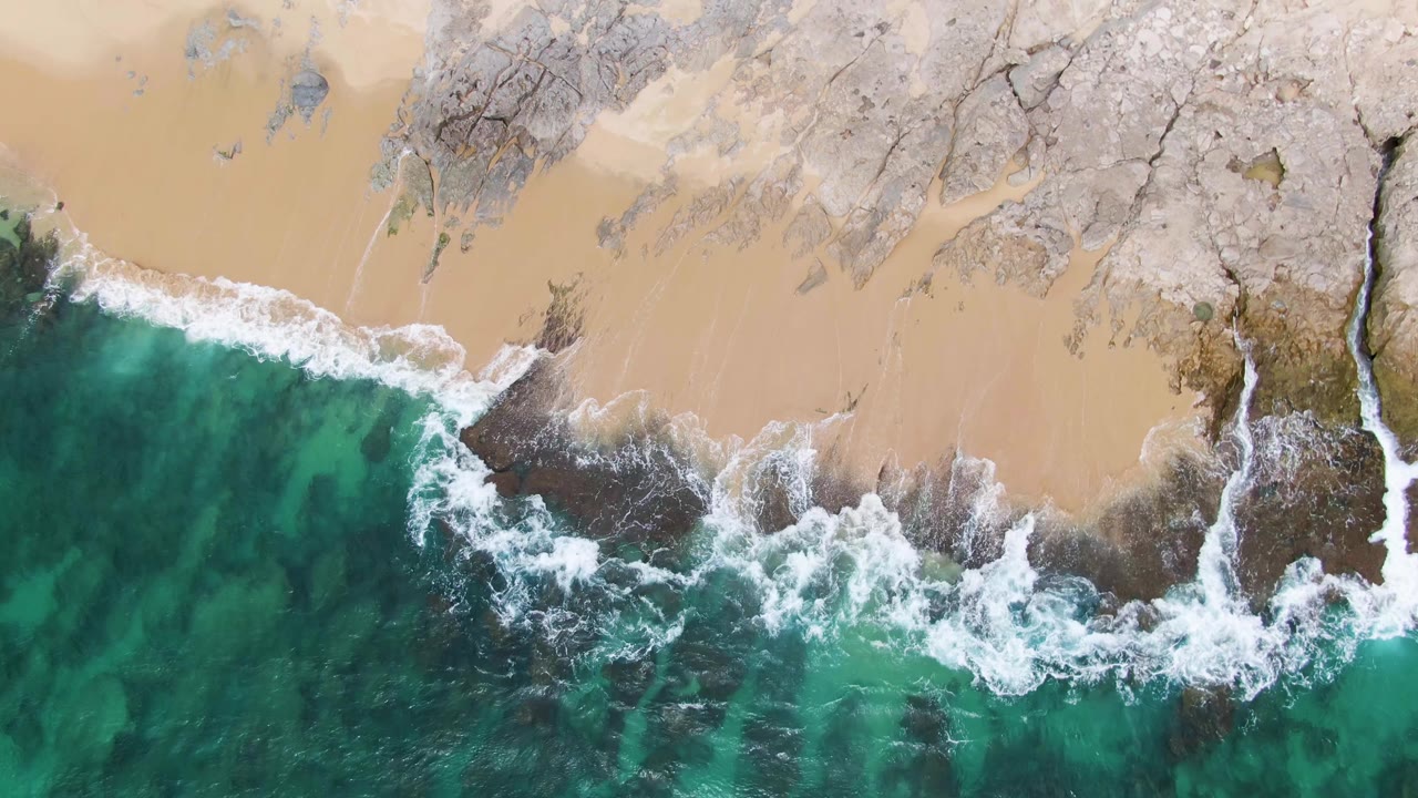 Top-down Punta Gorda Beach frame where white water breaks heavily over exposed dark rocks beside smooth sand and clear green-blue water, with no visible people.
