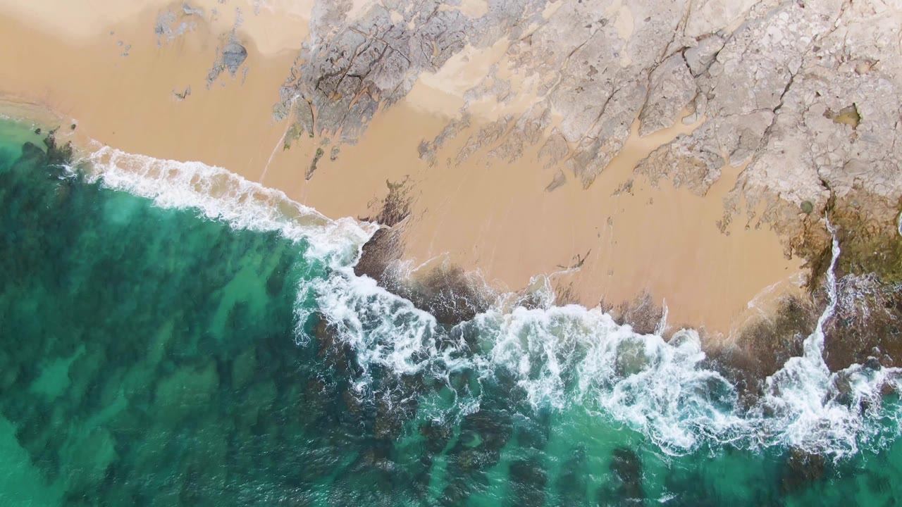 Punta Gorda Beach from above with water filling the lower frame, foam crossing the middle, and sandy beach with rock formations above, with no visible people.