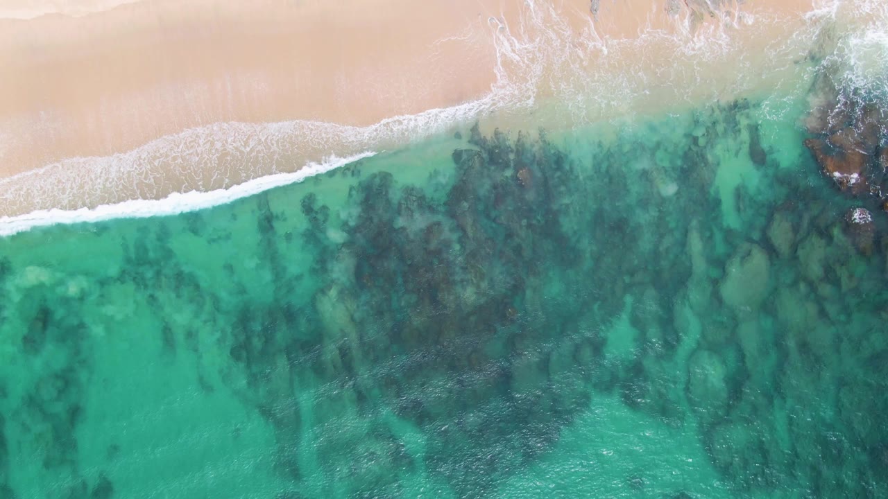 Overhead frame of Punta Gorda Beach with pale sand at the top, a narrow white surf line, turquoise water, dark seabed patches, and no visible people.