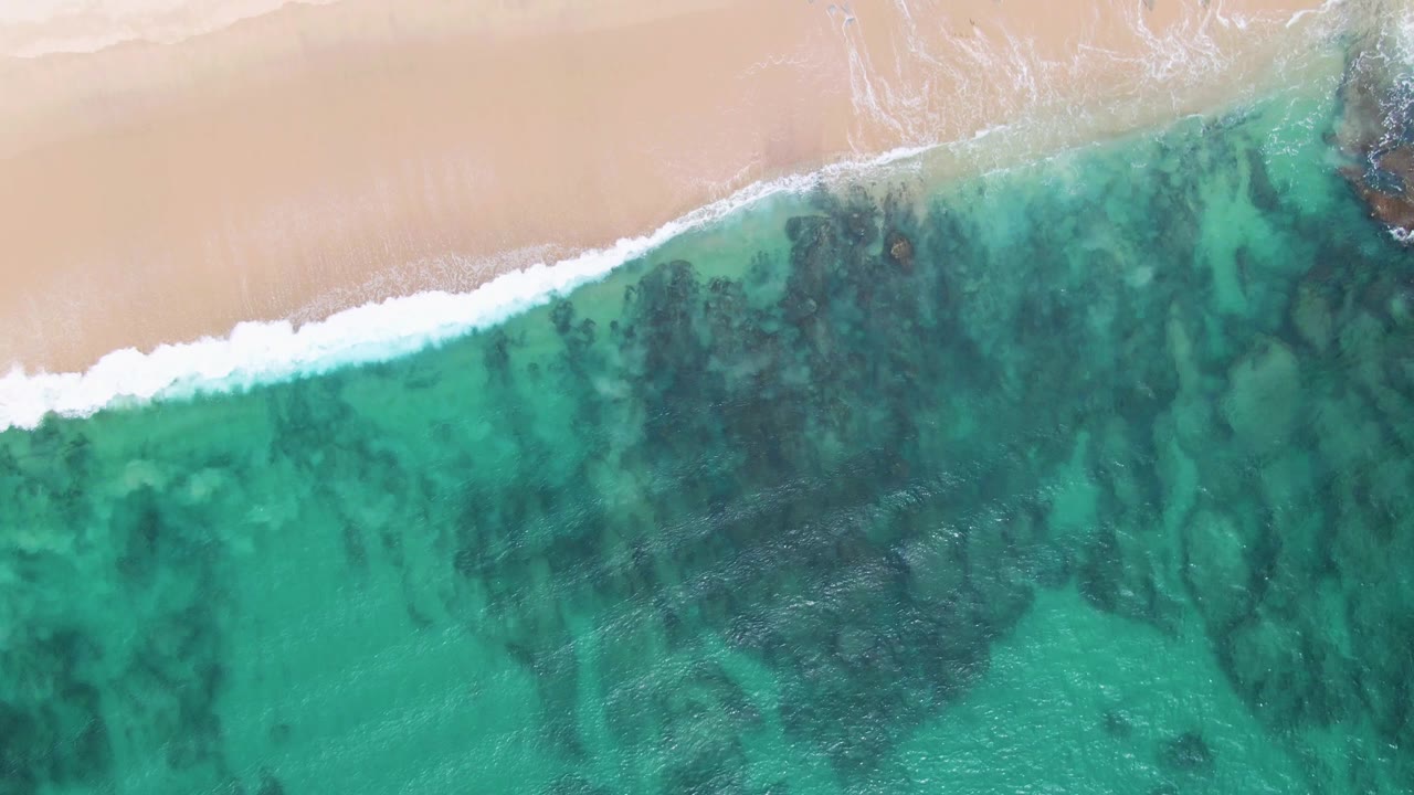 Aerial view at Punta Gorda Beach with the shoreline running diagonally, white foam breaking below pale sand, green-blue water below, and no visible people.