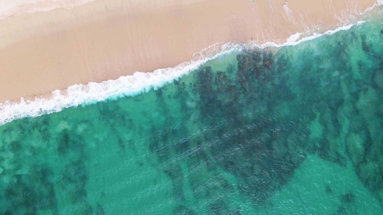 Top-down Punta Gorda Beach frame with a curved band of white foam along the sand and darker seabed patterns visible through the turquoise water, with no visible people.