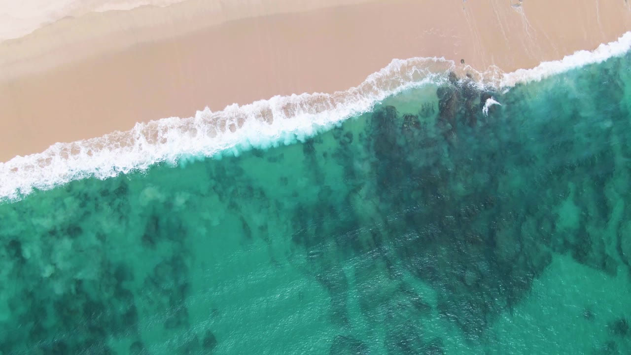 Aerial still from Punta Gorda Beach showing a wider white wave break across the upper water, pale sand beyond it, and clear turquoise shallows with no visible people.
