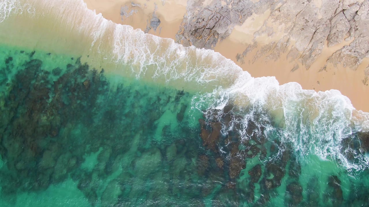 Top-down aerial frame at Punta Gorda Beach showing white surf over turquoise water, pale sand, dark underwater rocks, and no visible people.