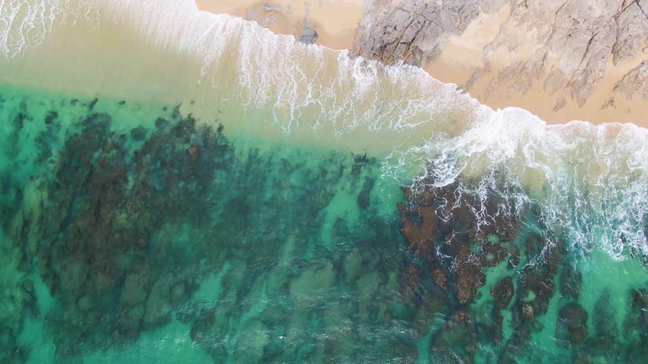 Aerial view at Punta Gorda Beach with a larger band of white foam pushing across dark submerged rocks and transparent turquoise water, with no visible people.