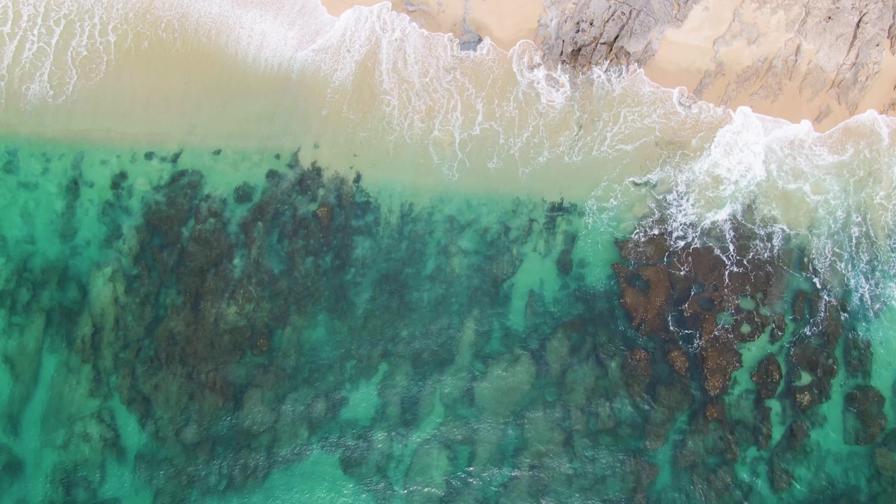 Overhead Punta Gorda Beach frame showing surf thinning along the sandy edge while clear green-blue water reveals dark seabed shapes and no visible people.