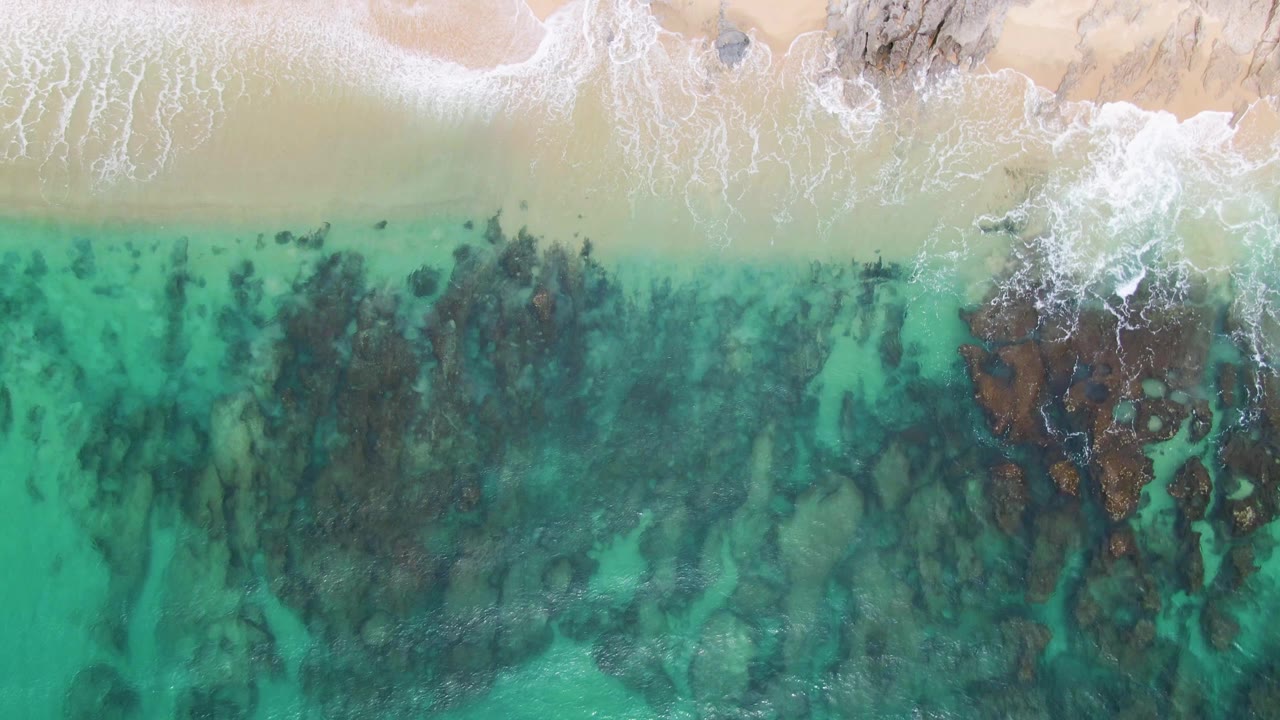 Aerial shoreline with foam crossing pale sand and turquoise shallows