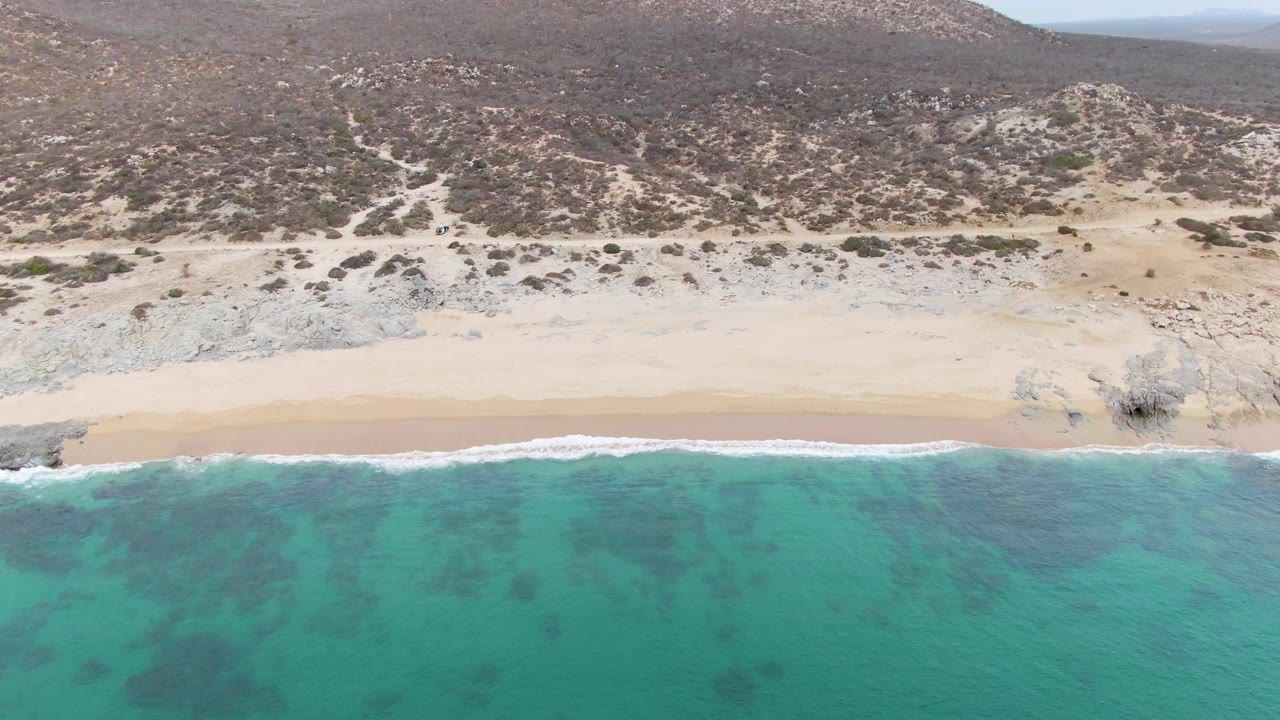 At Punta Gorda Beach in Baja California Sur, turquoise water meets a pale sandy shore below a dry rocky hillside with sparse shrubs and no visible people.