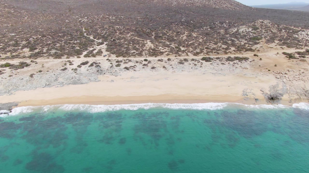 At 0:02, the aerial frame shows darker patches in the turquoise water near Punta Gorda Beach, a clean sand strip, and a rocky desert slope beyond, with no visible people.