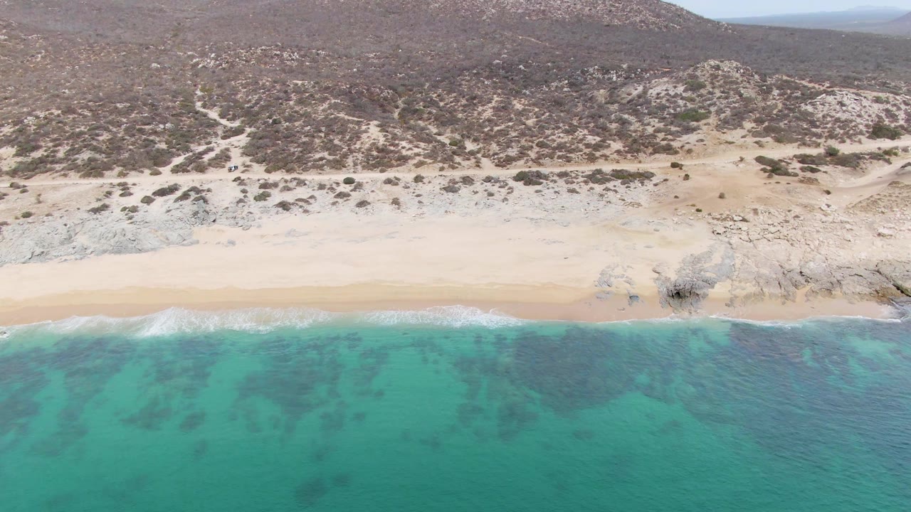 Aerial frame at Punta Gorda Beach, Baja California Sur, with clear turquoise water in front, foam sliding onto sand, scrub-covered hills behind, and no visible people.