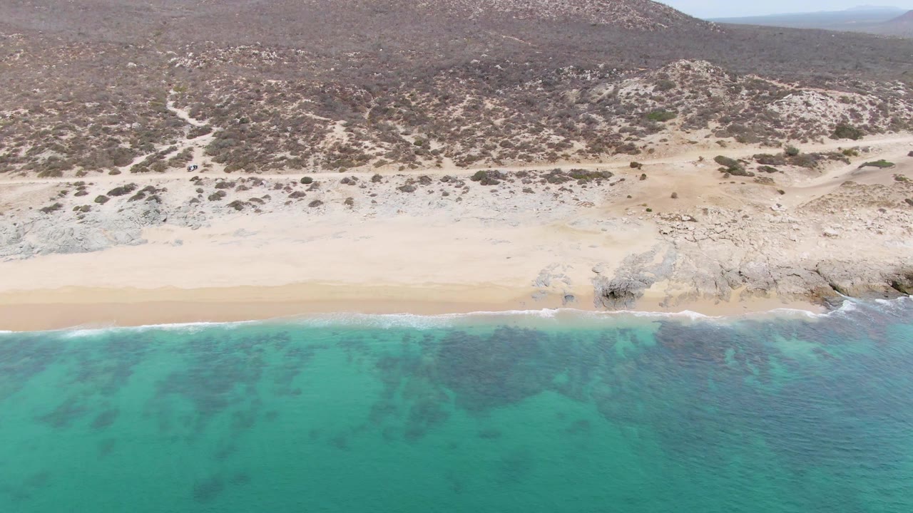 Aerial frame at Punta Gorda Beach, Baja California Sur, with turquoise water at the lower edge, white foam touching sand, rocky hillside above, and no visible people.