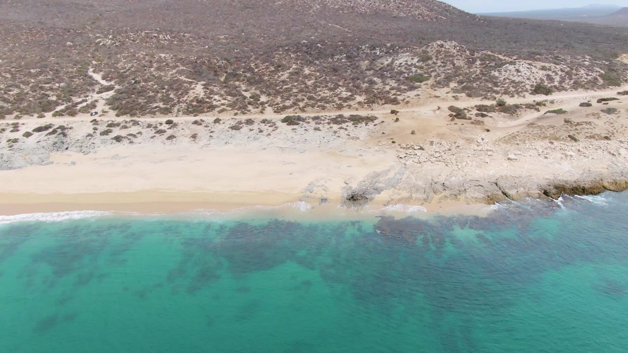 Aerial frame from Punta Gorda Beach showing turquoise water in the foreground, pale sand and rocks along the shore, dry hills behind, and no visible people.