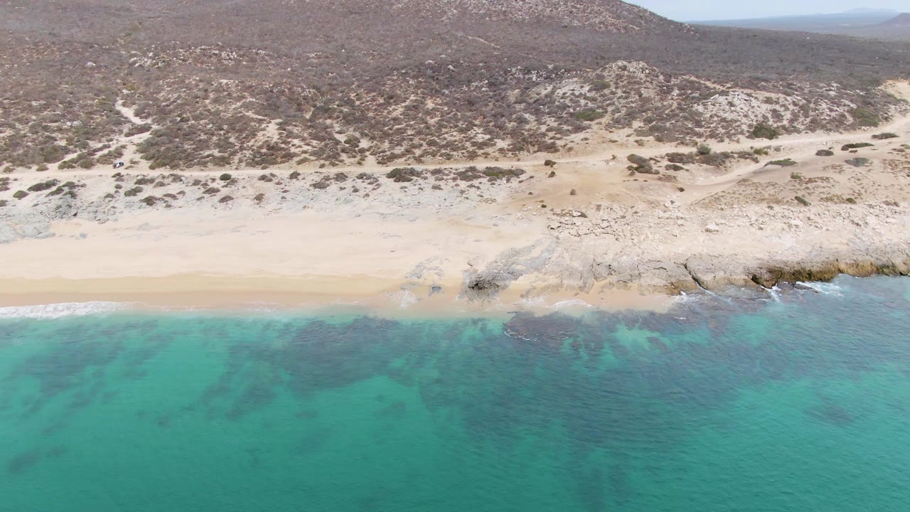 Punta Gorda Beach appears from above with a broad turquoise shallows area, a light sand strip, scattered shoreline rocks, scrubby hills, and no visible people.