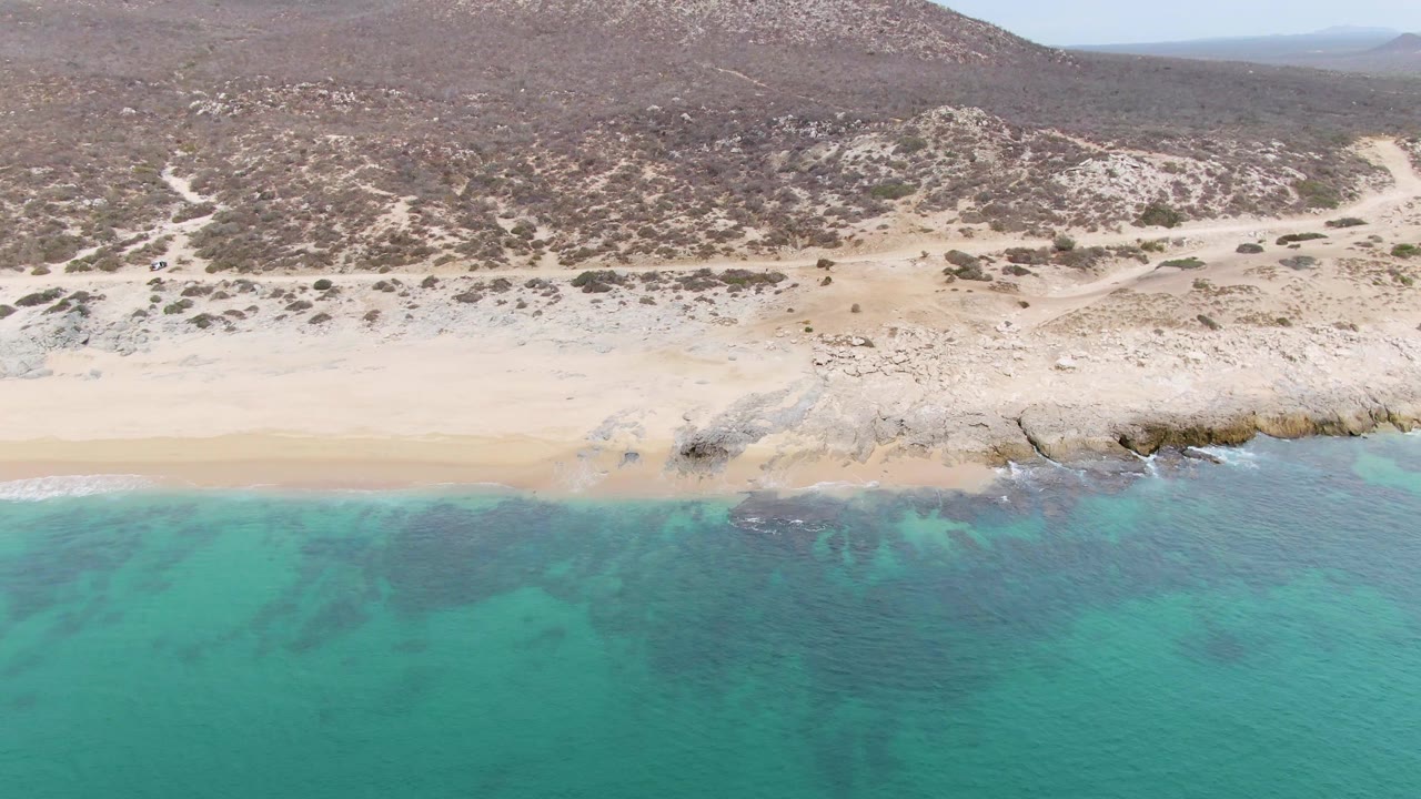 At 0:02, the Punta Gorda Beach shoreline shows clear turquoise water, darker submerged patches, pale sand, rocky outcrops, arid hills, and no visible people.