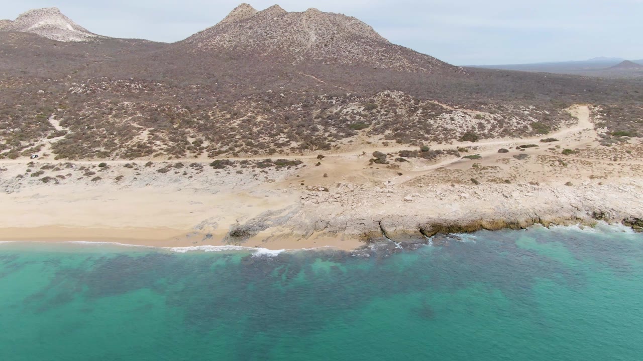 Wide aerial view of Punta Gorda Beach, Baja California Sur, with turquoise water in the foreground, a rocky sandy shoreline, desert scrub, mountains, and no visible people.