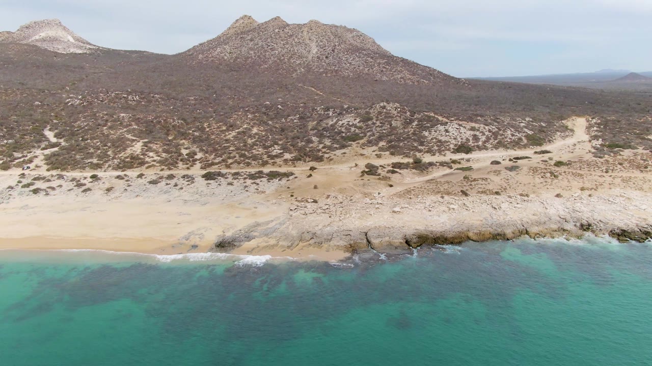 Aerial frame at Punta Gorda Beach with green-blue water, thin white foam along the shore, pale sand, rocky ledge, scrubby hills, mountains, and no visible people.