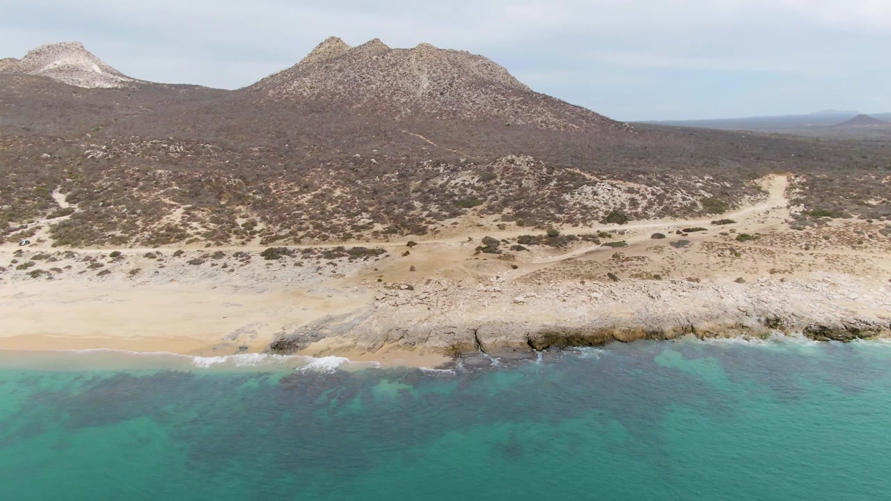 Punta Gorda Beach aerial with turquoise sea across the lower frame, broken rock along the shore, sandy beach, brown scrubland, distant mountains, and no visible people.