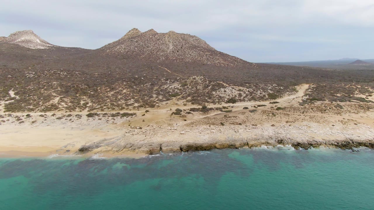 Aerial frame at Punta Gorda Beach, Baja California Sur, with turquoise water in the foreground, a pale rocky beach, scrub-covered desert, rugged mountains, and no visible people.