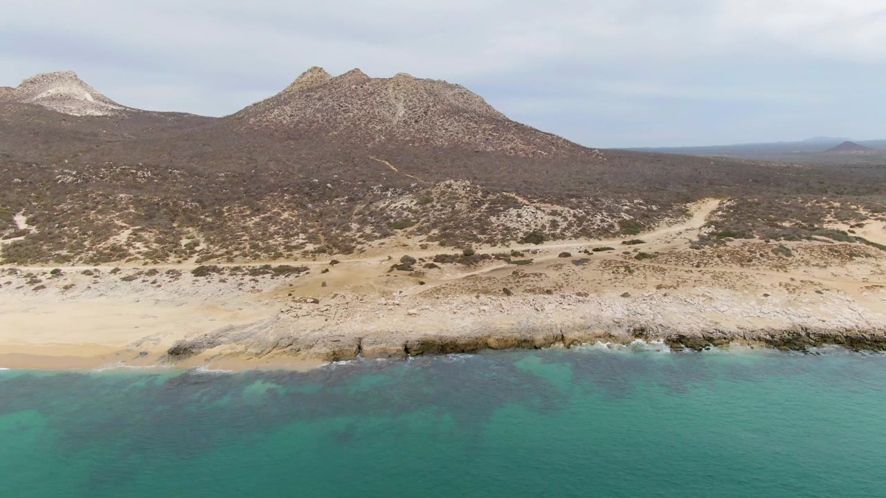 At 0:01, the Punta Gorda Beach aerial view holds on turquoise ocean, a narrow sandy and rocky shoreline, brown scrubland, distant mountains, and no visible people.