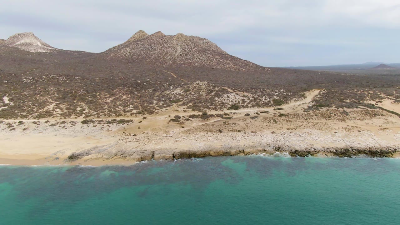 At 0:02, calm turquoise water sits below a pale Punta Gorda Beach shoreline, with dark desert scrub, rough mountain slopes, a cloudy sky, and no visible people.