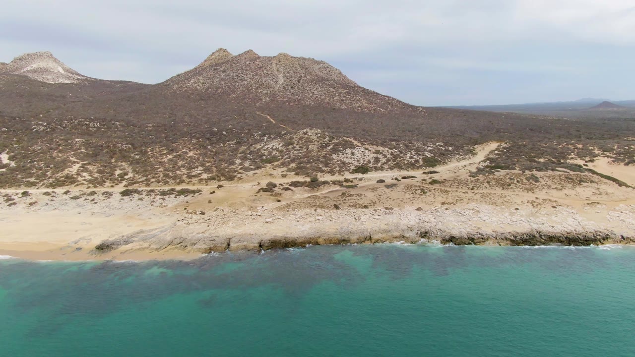 Aerial coastal landscape with waves, sandy beach, desert scrub, and rocky hills.