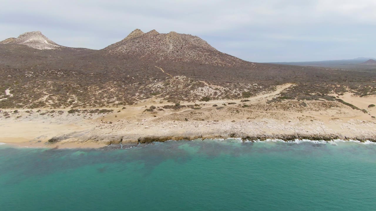 Aerial view at Punta Gorda Beach, Baja California Sur, with turquoise water in the foreground, a rocky surf line, pale sand, dry hills, distant mountains, and no visible people.