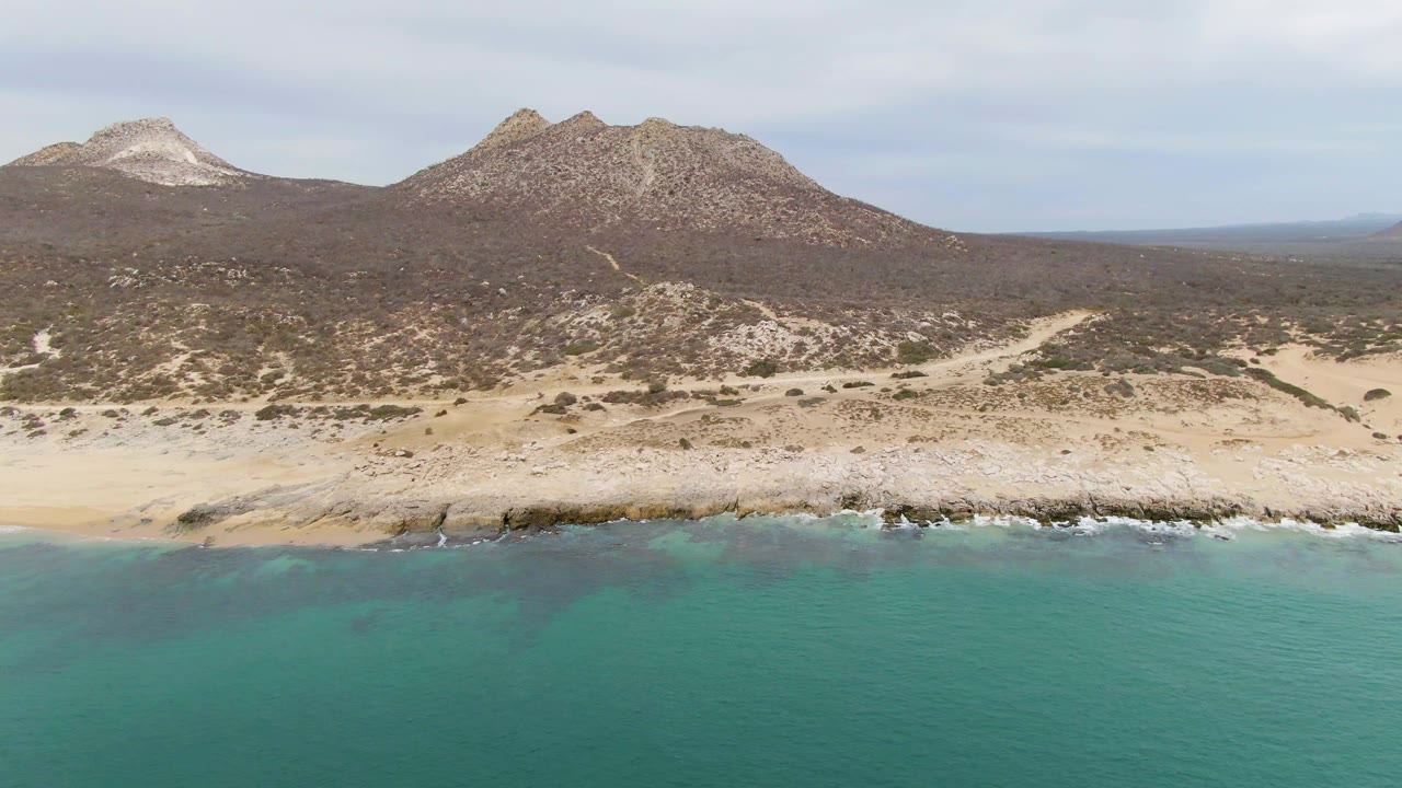 Punta Gorda Beach coastline seen from above the water, with a narrow sandy strip, dark rocks at the surf edge, scrub-covered desert terrain, mountains, and no visible people.