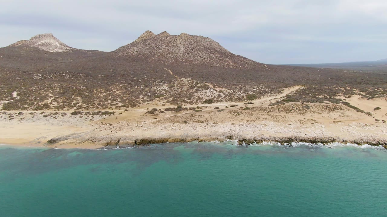 Wide aerial frame of Punta Gorda Beach with green-blue ocean across the lower half, light surf on the rocks, sandy shoreline, brown hills, distant peaks, and no visible people.