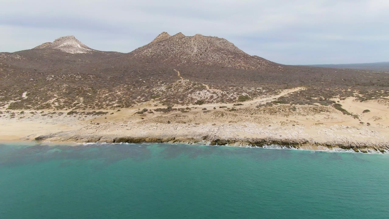 Wide aerial frame of Punta Gorda Beach in Baja California Sur with turquoise water in front, pale rocks at the shore, dry hills beyond, and no visible people.