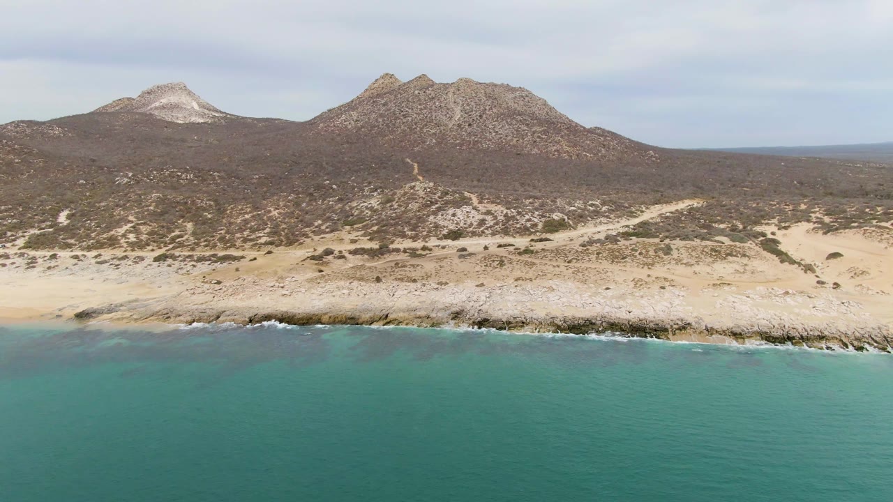 Aerial view at Punta Gorda Beach with a narrow white wave line along the rocks, turquoise water in the foreground, dry inland terrain, and no visible people.