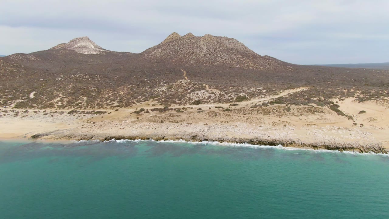 Punta Gorda Beach aerial frame with calm blue-green water below, a broken rocky shoreline across the middle, scrub-covered desert slopes, and no visible people.