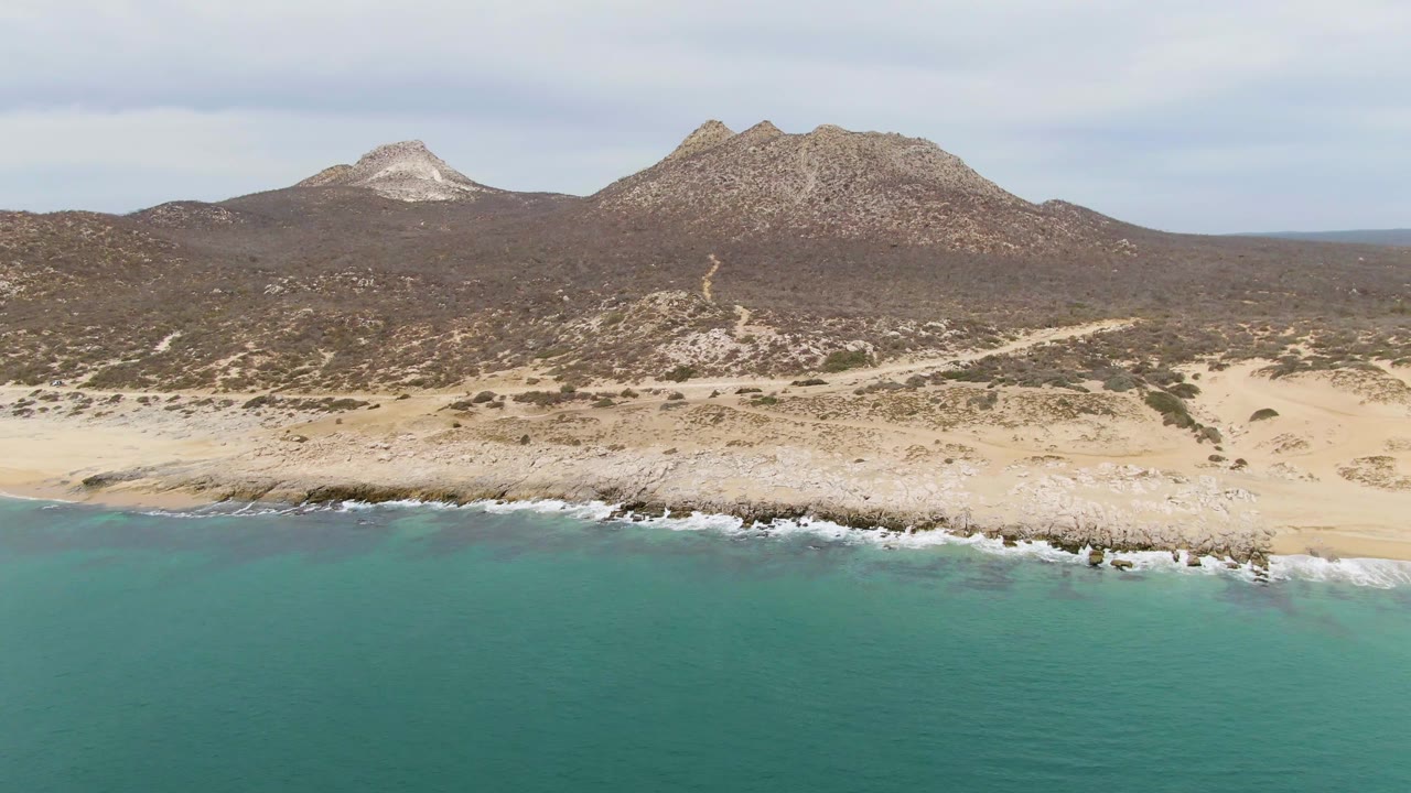 At Punta Gorda Beach in Baja California Sur, turquoise water meets a pale rocky shoreline with desert hills and two mountain forms in the background, with no visible people.