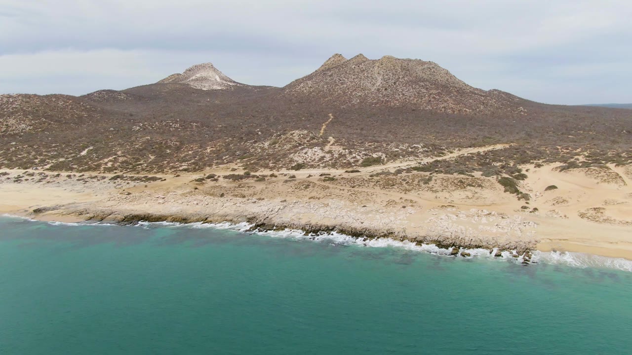 At Punta Gorda Beach in Baja California Sur, white water gathers against the rocks as the sandy beach and scrub-covered hills lead back to rugged mountains, with no visible people.