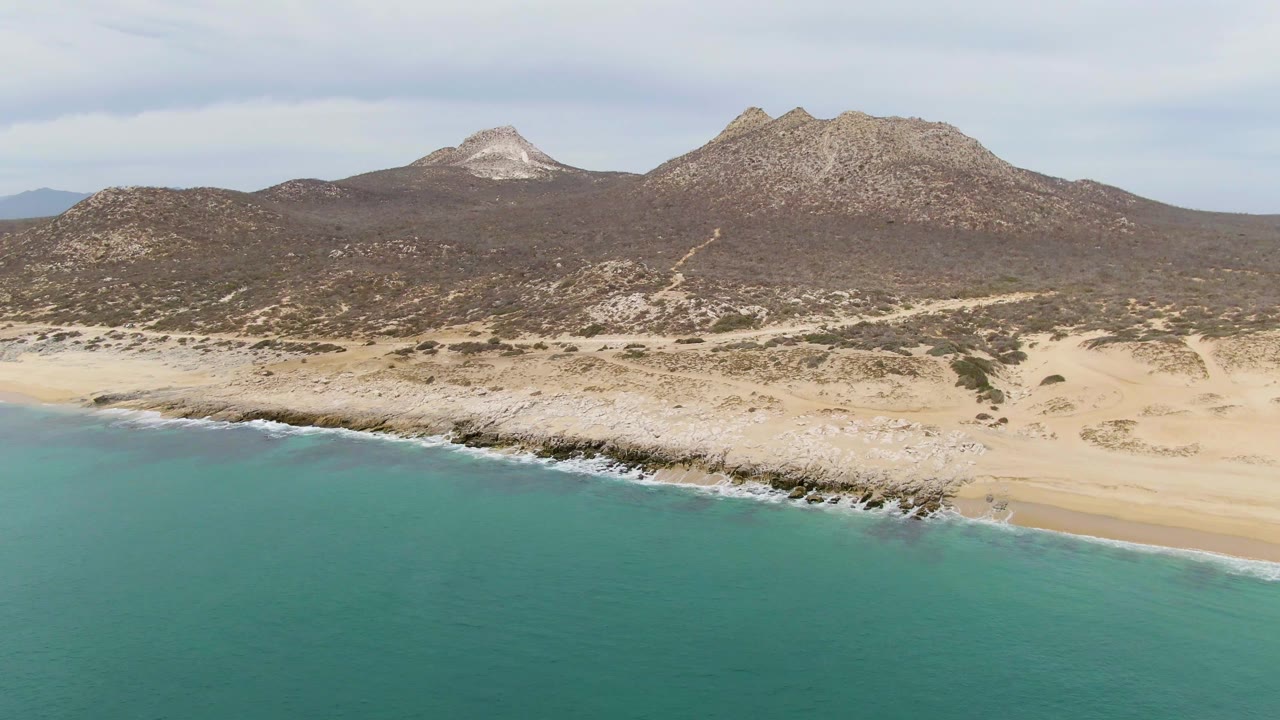 Opening frame of Punta Gorda Beach in Baja California Sur with turquoise ocean in the foreground, a sandy rocky shoreline, distant dry hills, and no visible people.