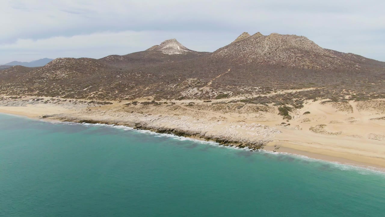 Mid-clip view of Punta Gorda Beach with more open water in the foreground, a pale beach and rock band across the center, dry mountain slopes, and no visible people.