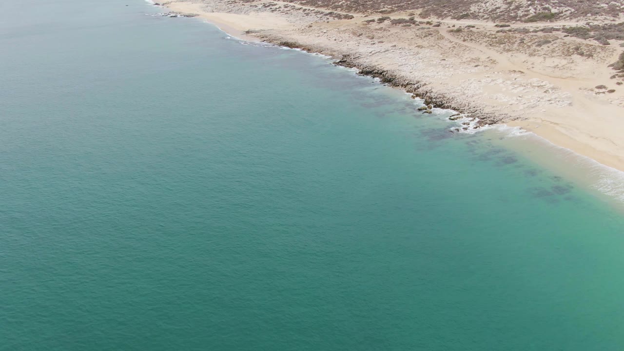 Aerial view at Punta Gorda Beach, Baja California Sur, with turquoise ocean beside pale sand, dark shoreline rocks, low scrub inland, and no visible people.