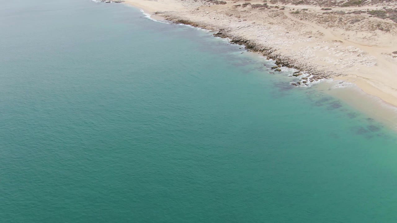 Aerial view over Punta Gorda Beach with clear green-blue water, a narrow line of surf, rocky patches near shore, scrubby dunes, and no visible people.