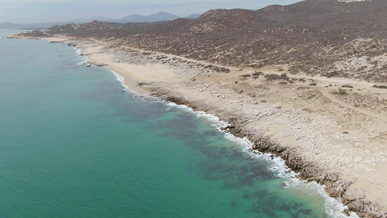 Aerial view of turquoise ocean beside a sandy rocky coastline and dry hills.