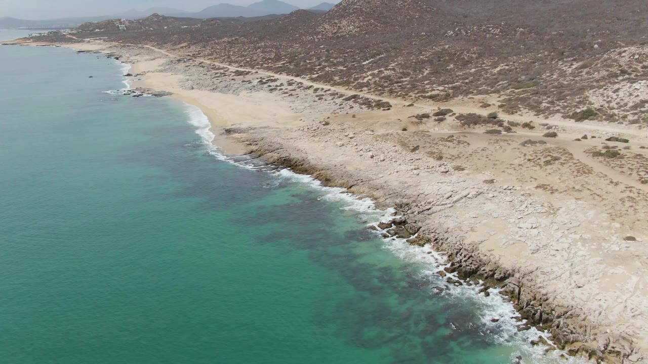 Aerial view over Punta Gorda Beach with the sea occupying the left frame, a curving rocky beach through the center, dry hills across the top, and no visible people.
